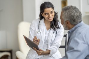 A young female doctor of Middle Eastern decent, sits with a senior patient in the comfort of his own home during a Homecare visit. She is wearing a white lab coat, has a stethoscope around her neck and is holding out a tablet as she shows the gentleman his recent test results. The senior is leaning in to look at the screen as he listens attentively.